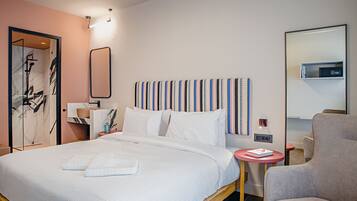 Une chambre d’hôtel moderne avec un grand lit, une table de chevet avec un livre, une chaise et une salle de bain équipée d’une douche et d’un miroir.