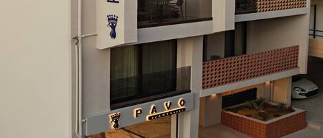 A multi-story building with balconies, a visible sign reading "PAVO," and a small outdoor seating area with plants.