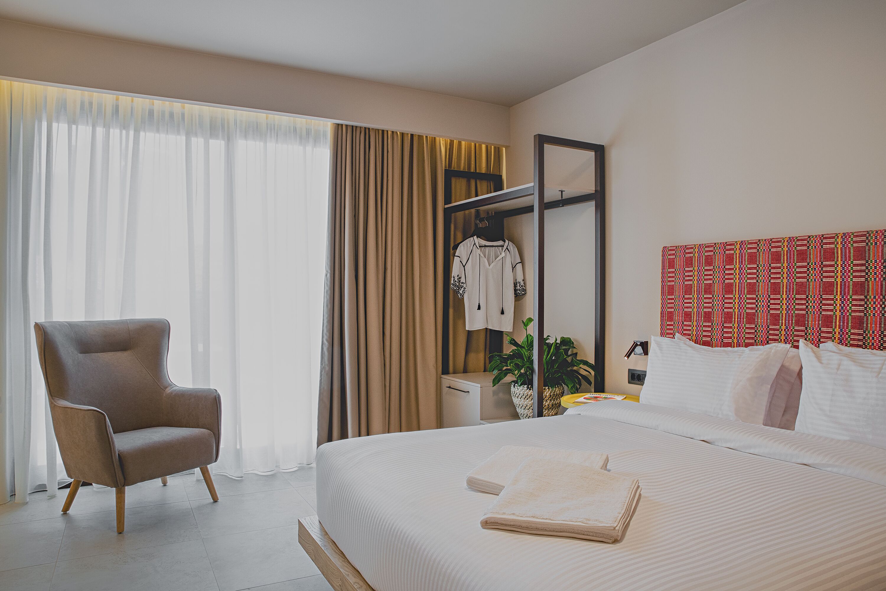 Een hotelkamer met een groot bed, een zithoek met een fauteuil, een kledingkast en een hoofdeinde met een rood patroon.