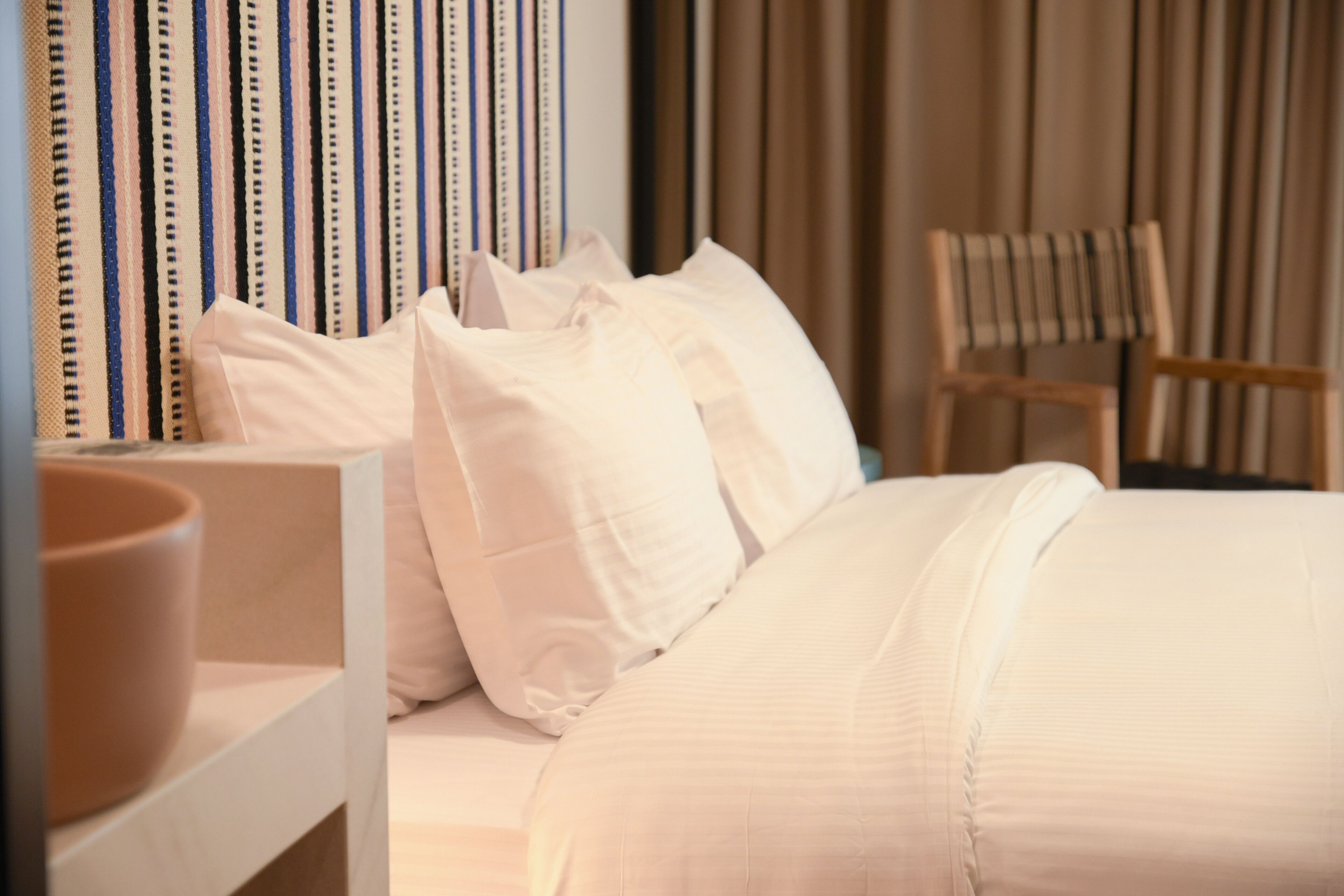 Ein ordentlich bezogenes Bett mit weißen Leinentüchern, ein Holzstuhl und gestreifte Vorhänge in einem Hotelzimmer.