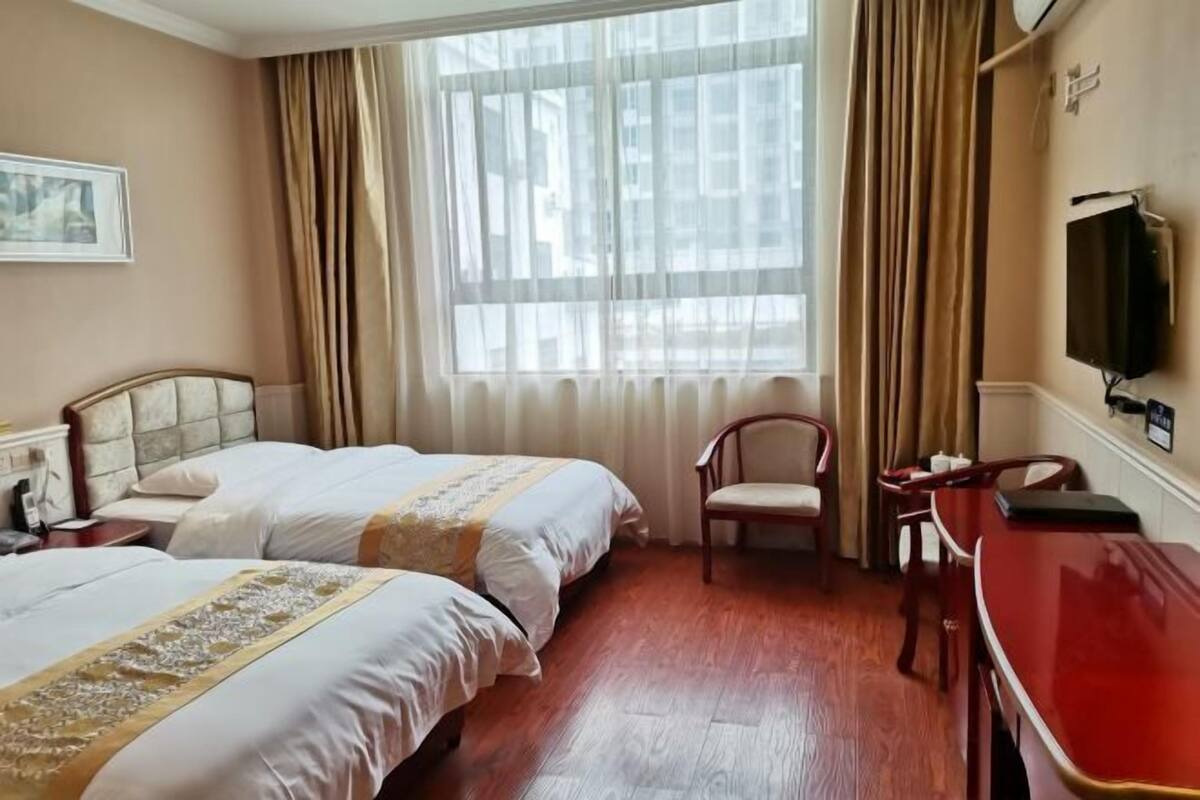 Une chambre d’hôtel avec deux lits, un bureau, une chaise, une télévision et une grande fenêtre avec des rideaux.