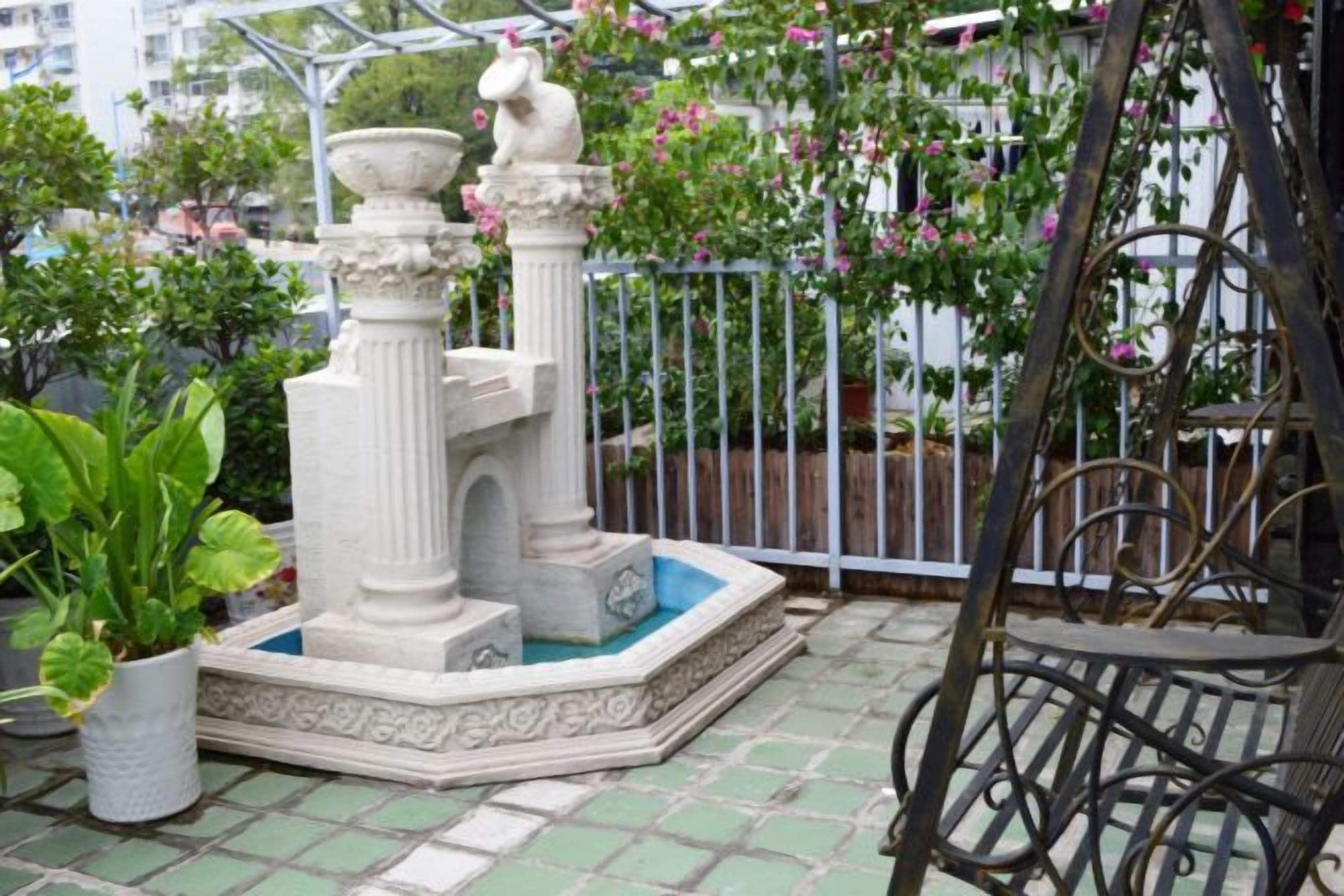 Um jardim com uma fonte, um banco de metal e uma cerca branca.