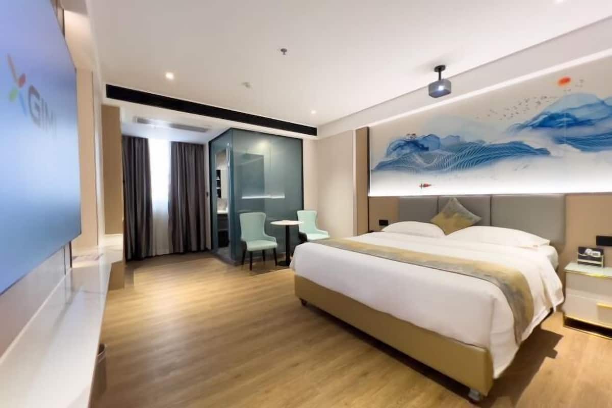 A modern hotel room with a large bed, a desk, and a chair. There is a painting on the wall and a TV on the left side.