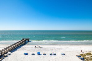 A beach with a wooden pier extending into the ocean, sandy beach, and several blue lounge chairs.