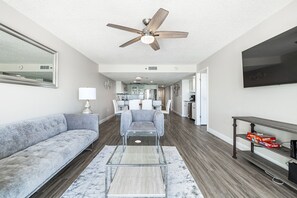 A spacious living room with a gray sofa, a coffee table, and a ceiling fan. A kitchen is visible in the background.