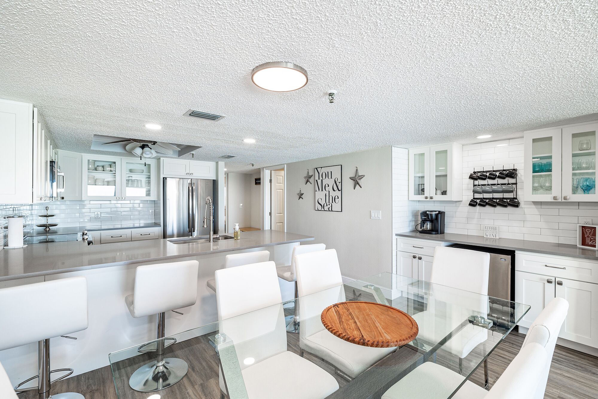 Una cocina moderna con muebles blancos, un refrigerador de acero inoxidable y una mesa de comedor de vidrio con sillas blancas.