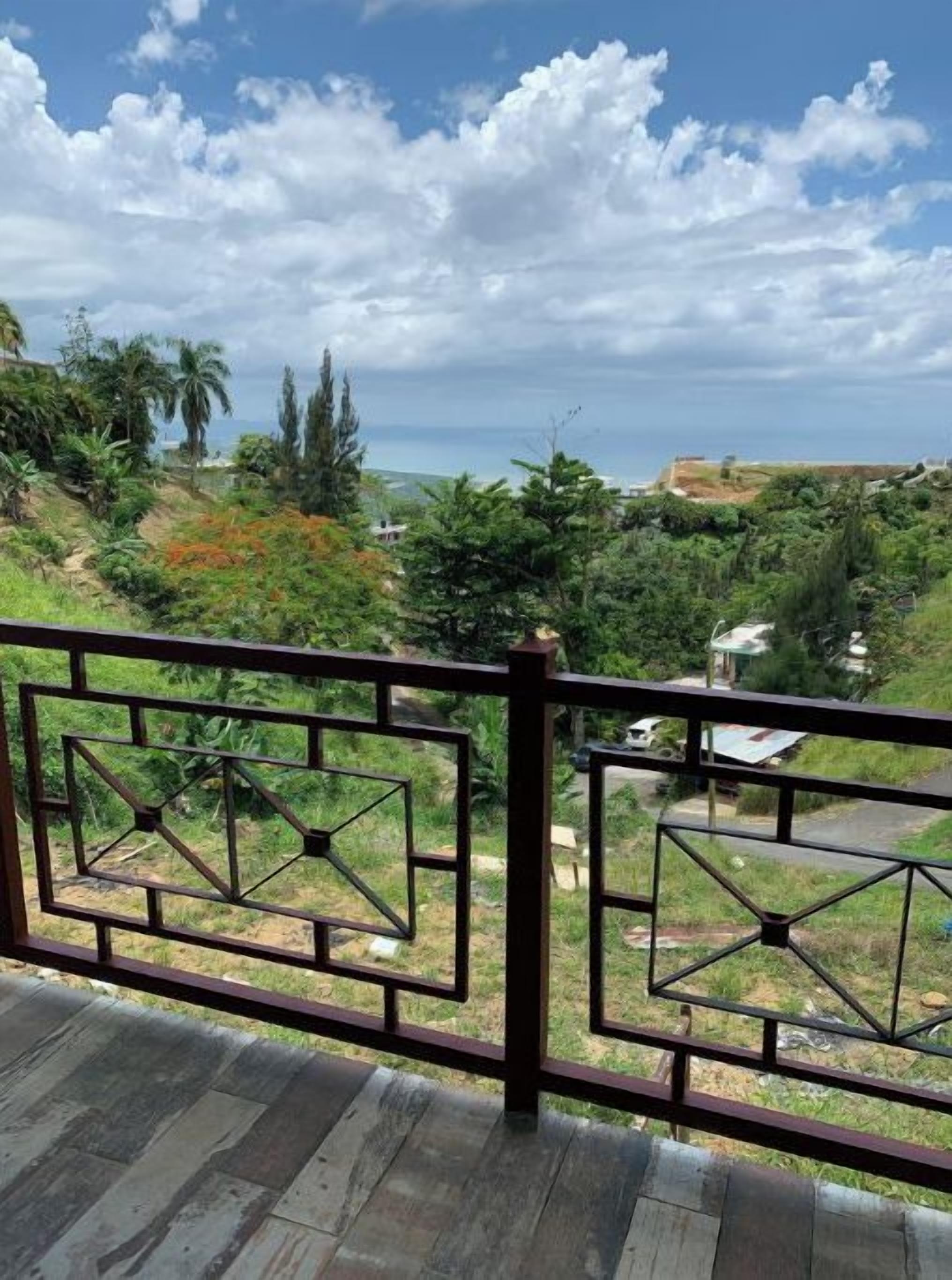 Un balcon doté d’une balustrade en métal, qui offre une vue sur un paysage luxuriant, avec des arbres et, au loin, la mer.