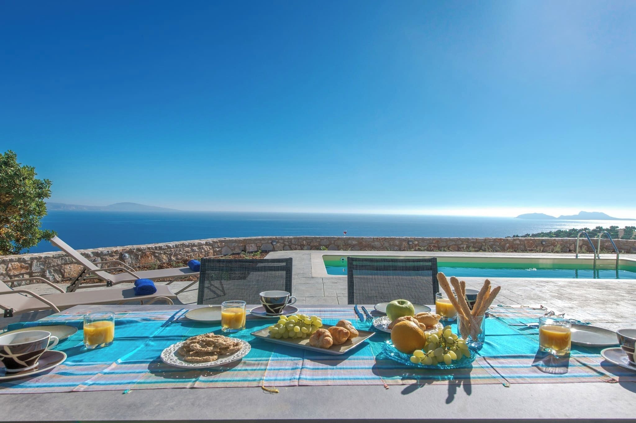 A breakfast setup with a view of the sea, including plates of food, glasses of juice, and a table with a blue tablecloth.