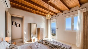 A bedroom with a wooden ceiling, a large bed with white linens, a bedside table with a lamp, a wooden wardrobe, and a bathroom visible through an open door. - Tranquil sea view villa with pool,2km from beach! (Agia Galini)