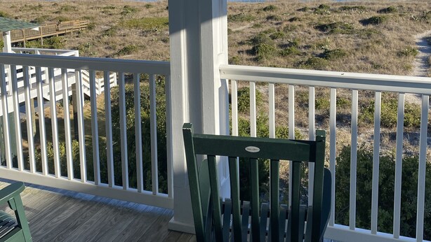 A wooden deck with a white railing, a green chair, and a view of the beach and ocean.