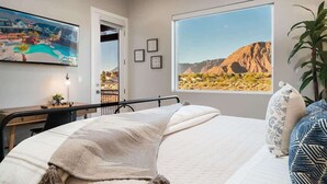 Un dormitorio con cama, escritorio, silla, ventana con vista al desierto y cuadros en la pared.