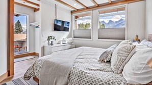 A bedroom with a bed, a TV, a desk, and a window with a mountain view. - Raven's Nest - Perfectly Located on Main Street (Canmore)