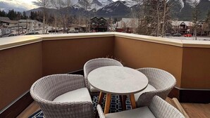 A balcony with wicker chairs and a table, overlooking a mountainous landscape with snow-capped peaks. - Raven's Nest - Perfectly Located on Main Street (Canmore)