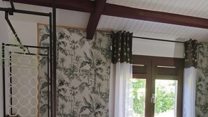 A room with a tiled floor, a window with white curtains, a wall with a leaf pattern, and a shelf with leopard print pillows. - LES FLAMBOYANTS LA PETITE MAISON CRÉOLE (Anse-Bertrand)