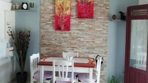A dining area with a stone wall, two paintings, a white table with chairs, and a potted plant. - LES FLAMBOYANTS LA PETITE MAISON CRÉOLE (Anse-Bertrand)