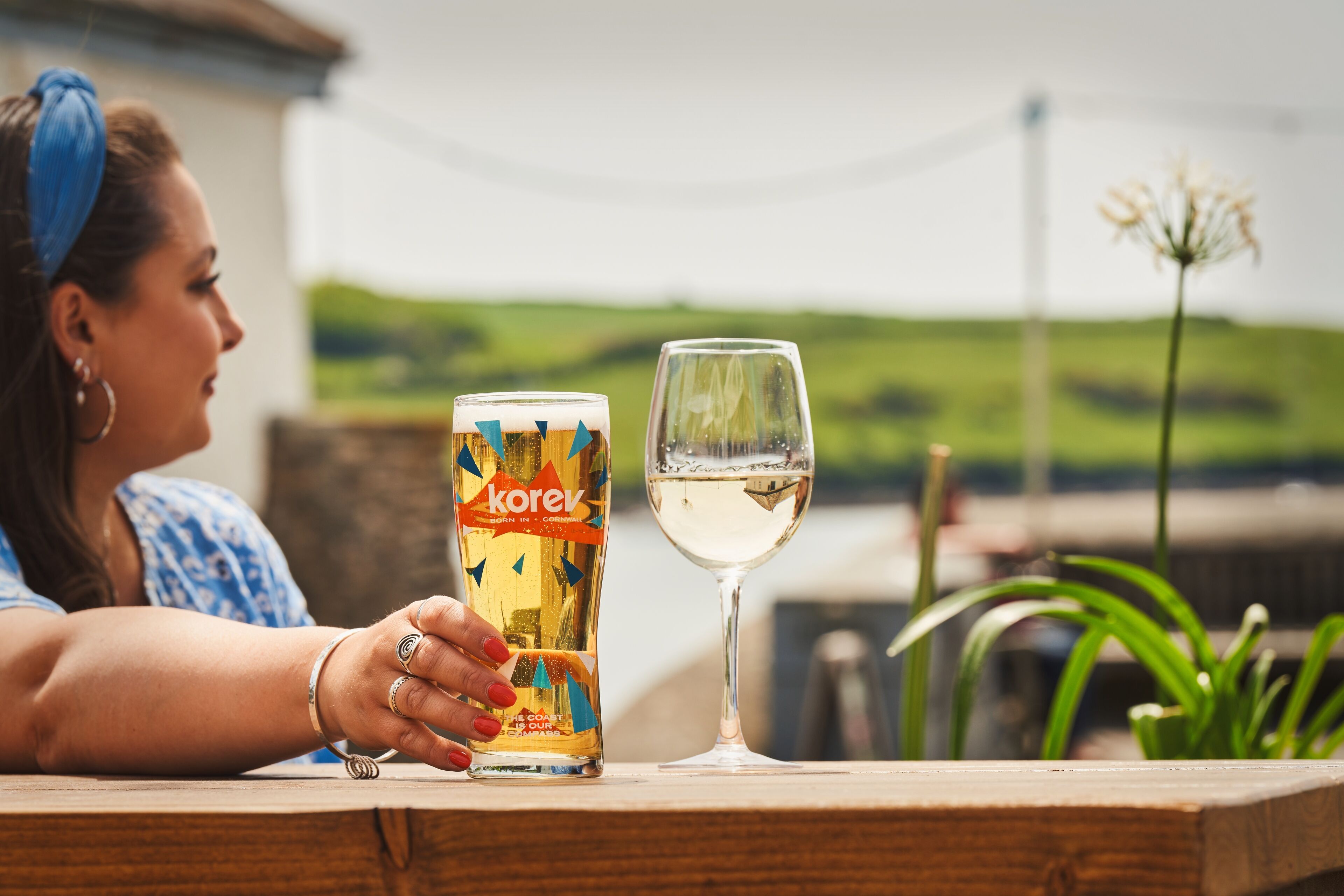 A person sitting at a wooden table outdoors with a bottle of Korey beer and a glass of white wine.