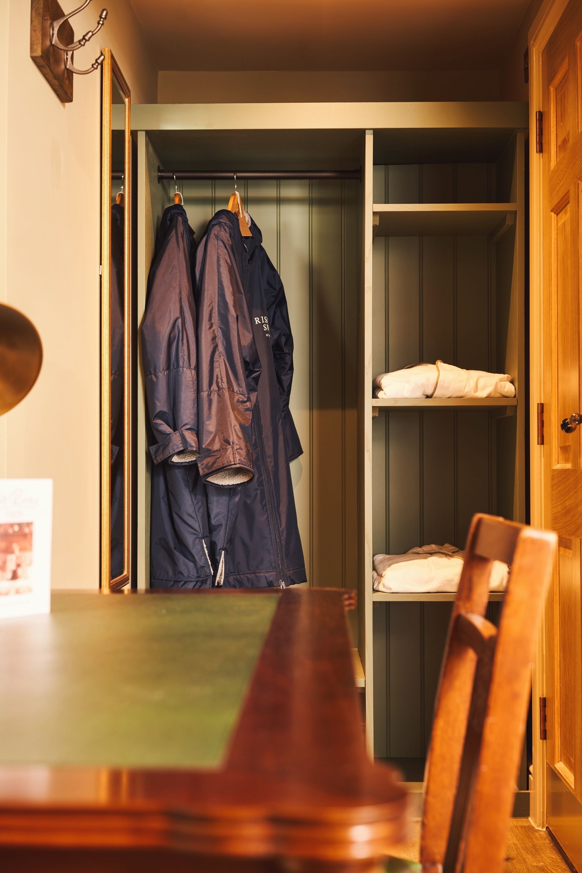 A room with a wooden dining table, a coat rack with two jackets, and a wooden wardrobe with a folded towel.