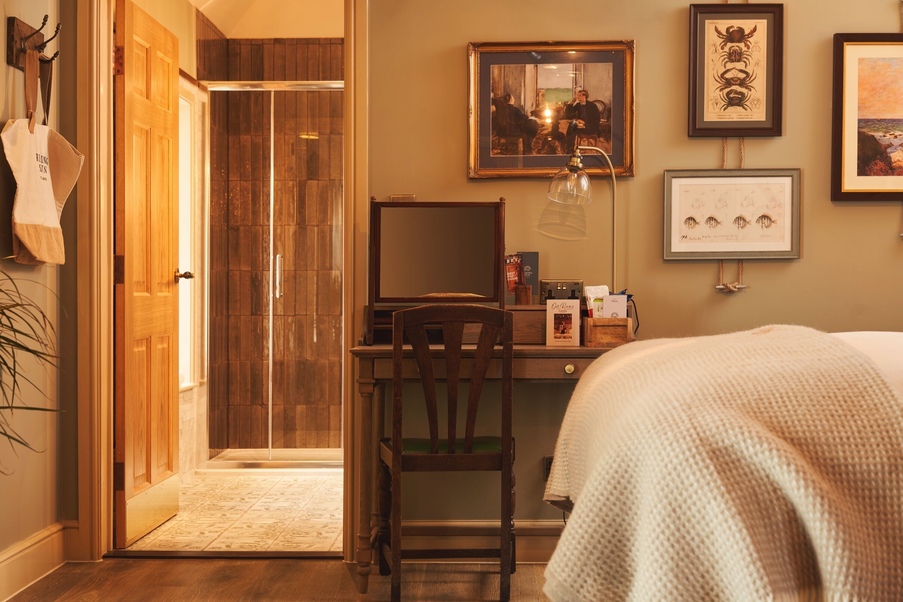 Quarto de hotel com escrivaninha de madeira, uma cadeira, cama com cobertura branca, área de chuveiro e quadros na parede.