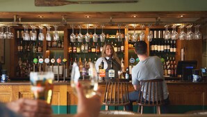 Un bar avec des étagères en bois remplies de bouteilles, un barman et des clients tenant des verres.