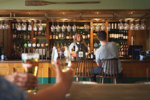 Een bar met houten planken vol flessen, een barman en gasten met glazen.
