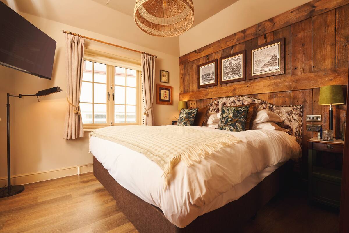 Quarto com cama grande, cabeceira de madeira, quadros emoldurados, janela com cortinas e luminária de chão.