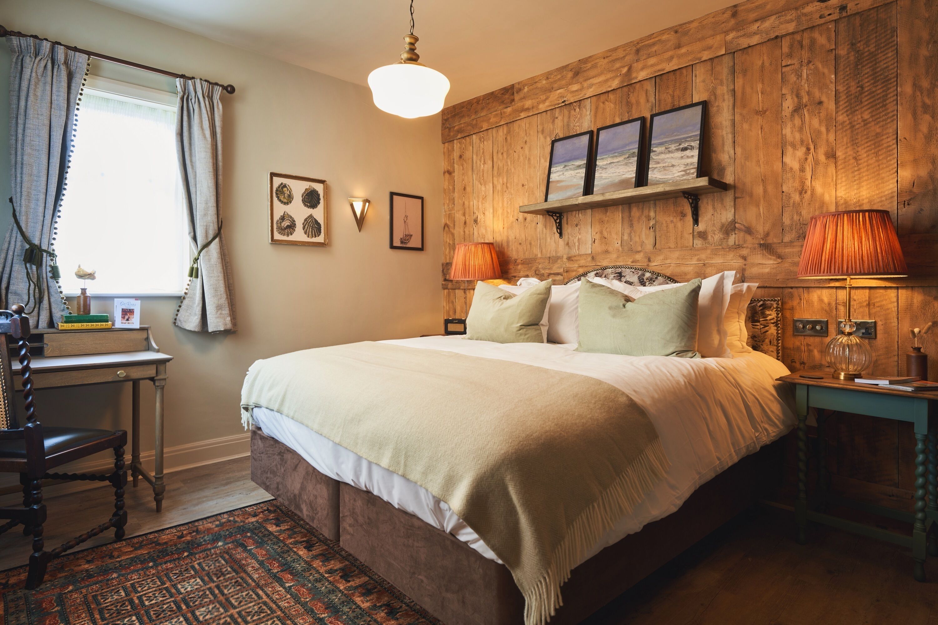 A bedroom with a wooden headboard, a bed with multiple pillows, a bedside table with a lamp, and a window with curtains.