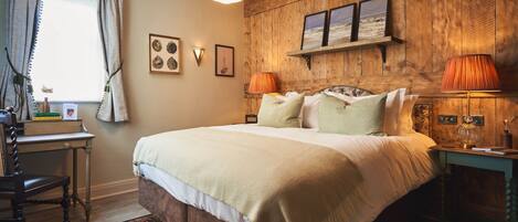 Un dormitorio con cabecera de madera, cama con varias almohadas, mesita de noche con lámpara y ventana con cortinas.