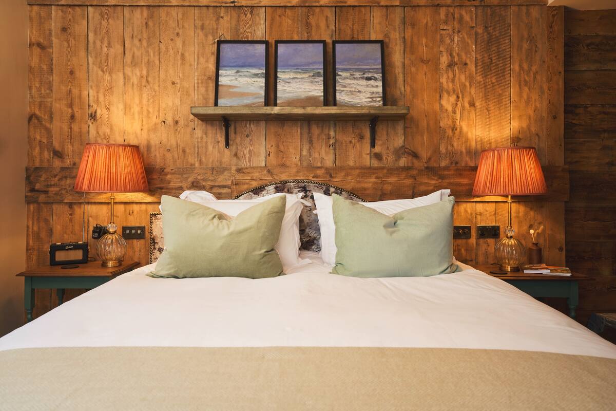 Quarto com cabeceira de madeira, cama com roupa de cama branca, duas almofadas verdes e duas luminárias de mesa.