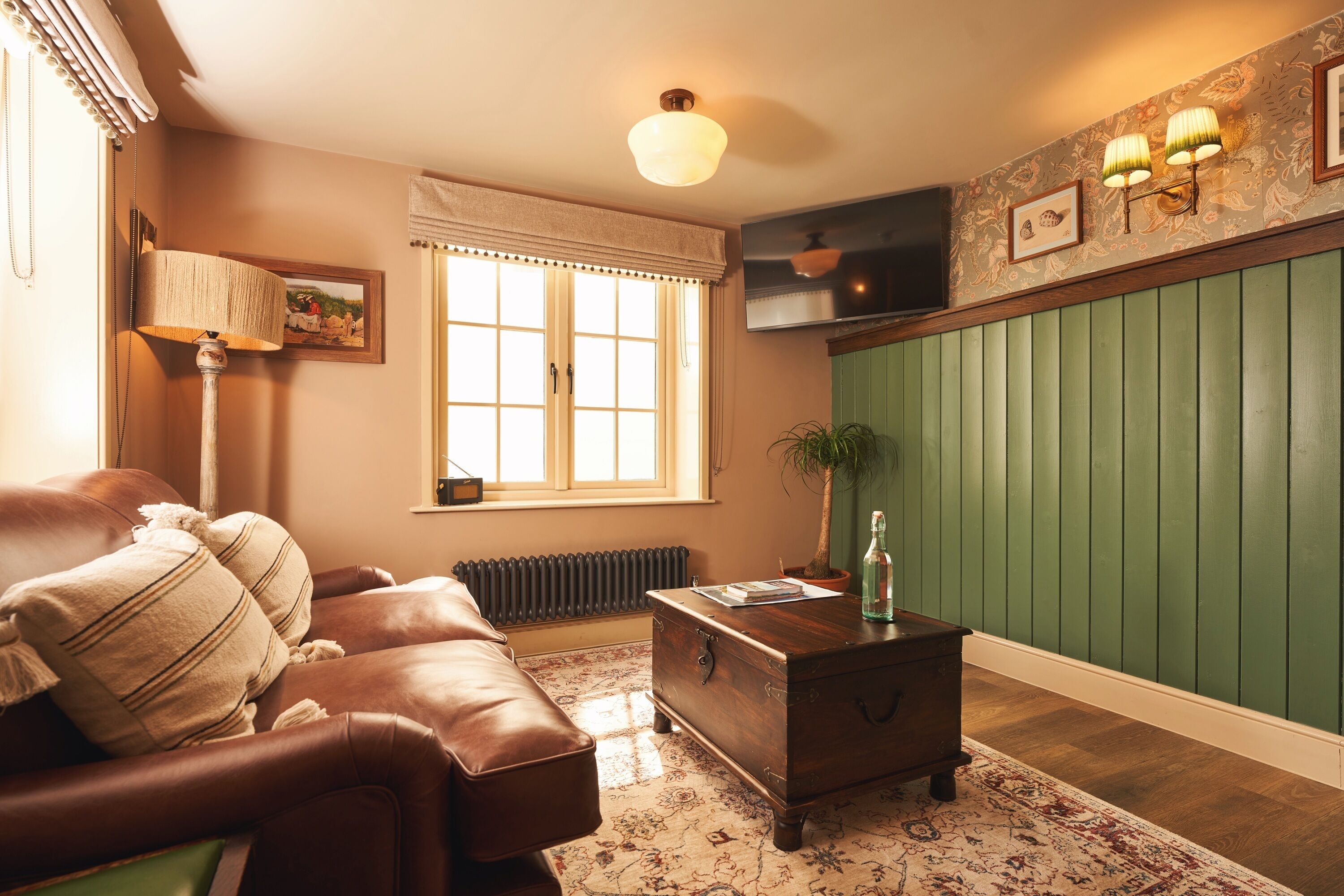 Una sala de estar con un sofá de cuero marrón, una mesa de centro de madera, un radiador, una pared verde con paneles y una ventana con cortina blanca.