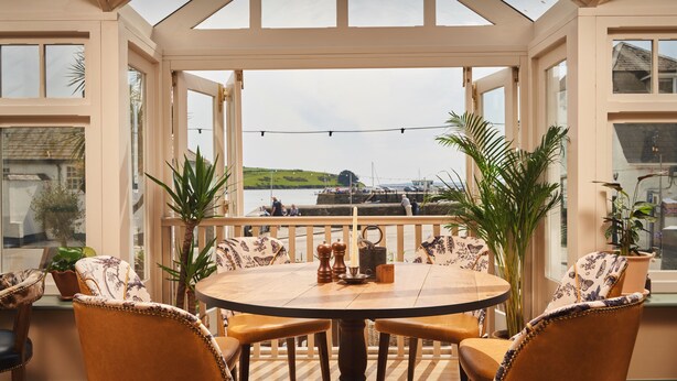 Une véranda avec une table et des chaises en bois, de grandes fenêtres et une vue sur la mer.