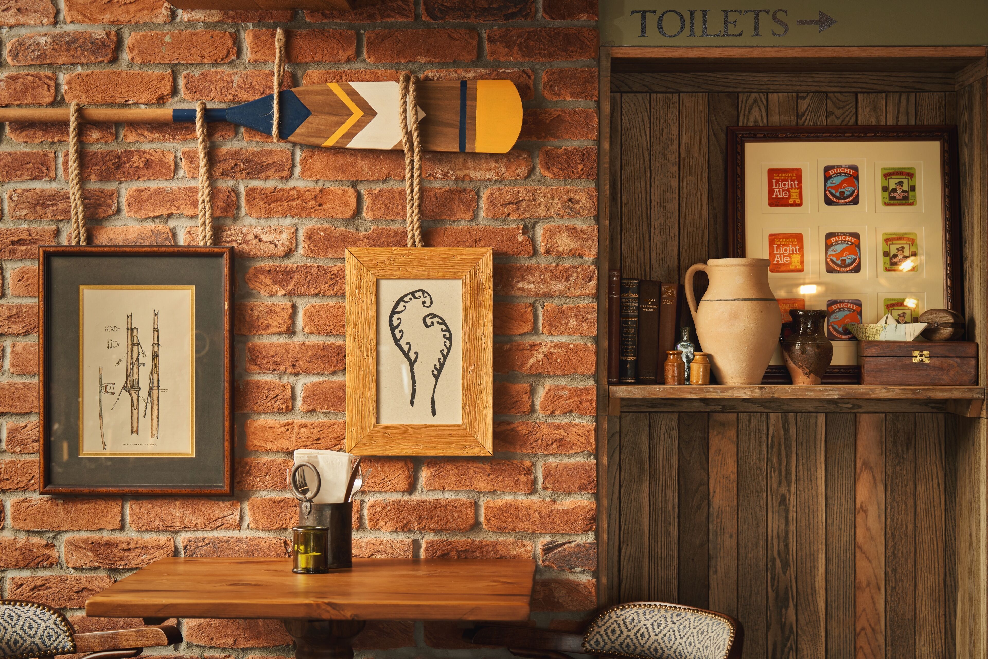 A rustic brick wall with framed artwork, a wooden table with a coffee cup, and a shelf with books and a vase.