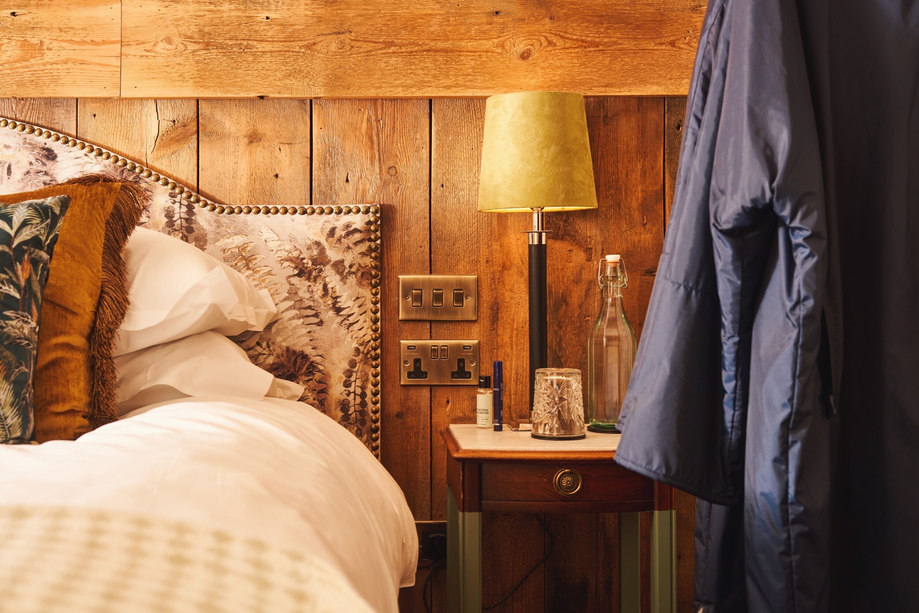 A wooden bedroom with a bed, a bedside table with a lamp, a bottle, and a glass container.