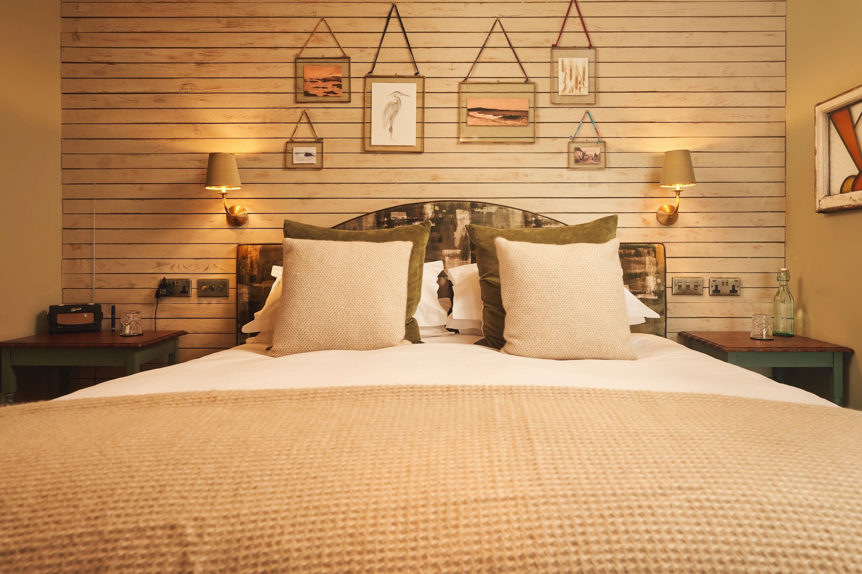 Une chambre avec un mur recouvert de lambris en bois, un lit avec une couette blanche et deux oreillers, des lampes de chevet et des œuvres d’art encadrées.