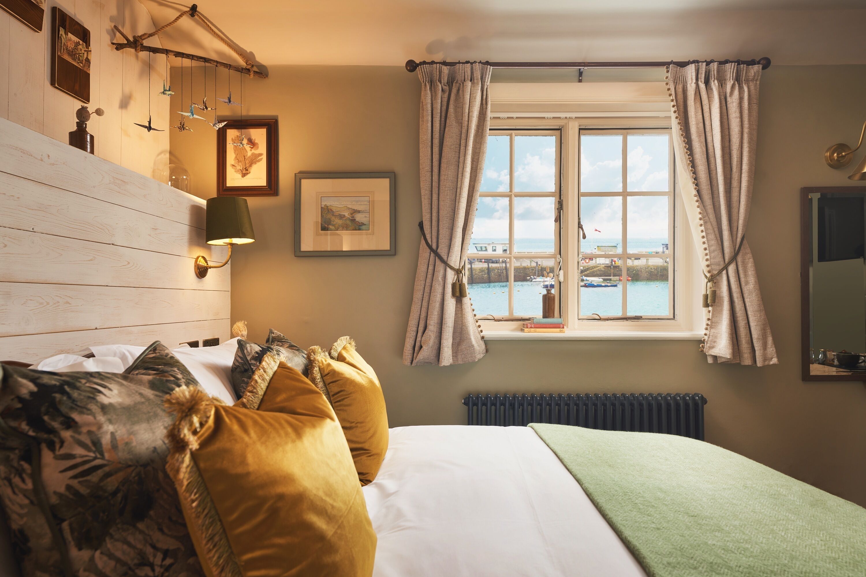 A bedroom with a wooden headboard, a bed with patterned pillows, a bedside lamp, a framed picture, a window with curtains, and a view of the sea.