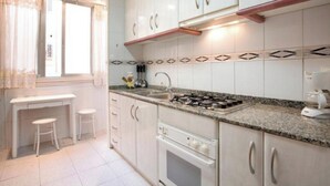 A kitchen with white cabinets, a gas stove, and a sink. There are two stools and a window with curtains. - Plaza Universitat Apartment (Barcelona)