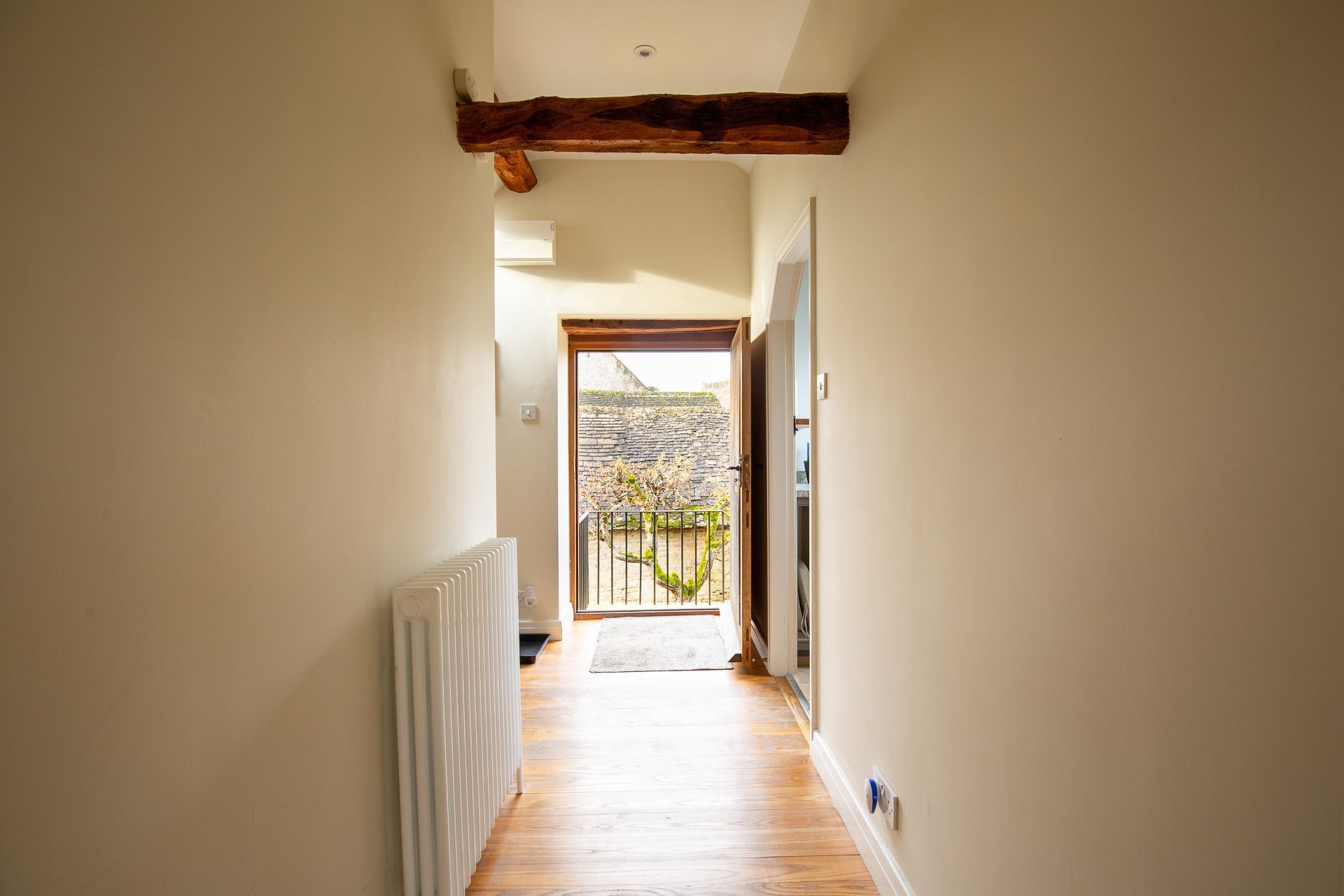 Un couloir étroit avec des poutres en bois au plafond, un radiateur sur le côté gauche et une porte au fond qui donne sur l’extérieur.