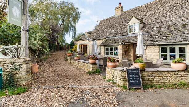 A traditional stone building with a signboard, outdoor seating area with potted plants, and a chalkboard displaying opening times.