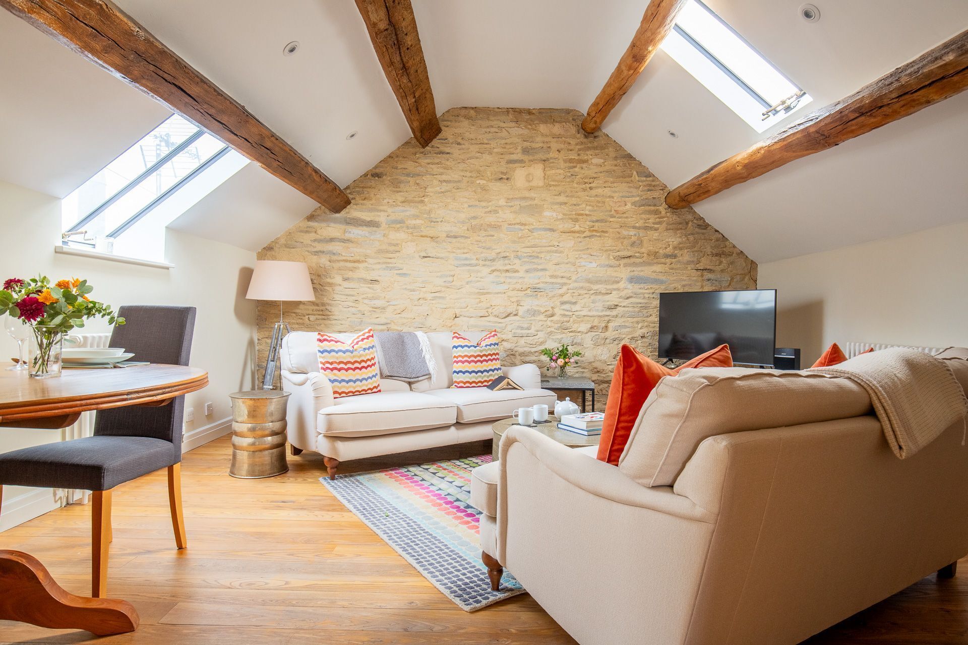 A cozy loft-style living room with exposed wooden beams, a stone wall, a white sofa, a TV, and a dining area with a table and chairs.
