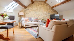 A cozy loft-style living room with exposed wooden beams, a stone wall, a white sofa, a TV, and a dining area with a table and chairs.