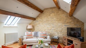 A cozy loft apartment with exposed wooden beams, a stone wall, a white sofa with colorful pillows, a coffee table with a vase of flowers, and a television on a wooden cabinet.