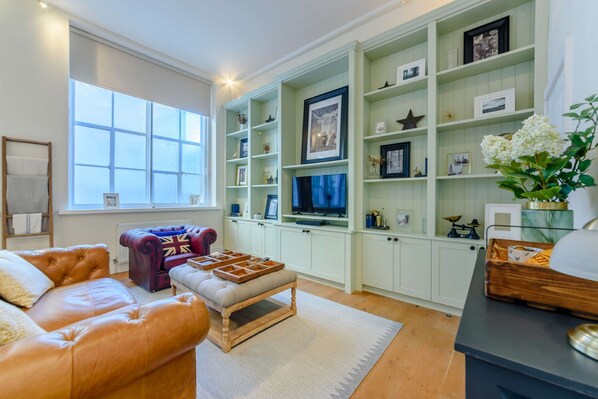 Ein Wohnzimmer mit einem Ledersofa, einem roten und blauen Sessel, einem Fernseher und einem Bücherregal mit verschiedenen Gegenständen.
