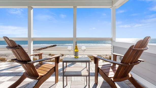 Un área de estar al aire libre cubierta con dos sillas de madera y una mesa, con vista a una playa.