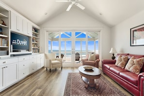 A spacious living room with a large window, a ceiling fan, a red leather sofa, a white armchair, a coffee table, and a bookshelf.