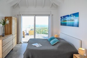 Relax in this serene bedroom with a large bed, dresser, and sliding door leading to a balcony with a coastal view.