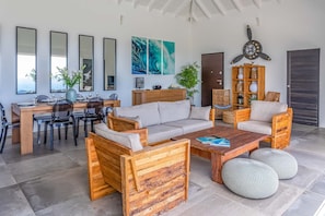 A modern living room with a sofa, coffee table, and chairs, a dining area with a table and chairs, and a kitchen area with a wooden cabinet and a clock. - Villa La Vue (1 bedroom) (Saint Barthélemy)