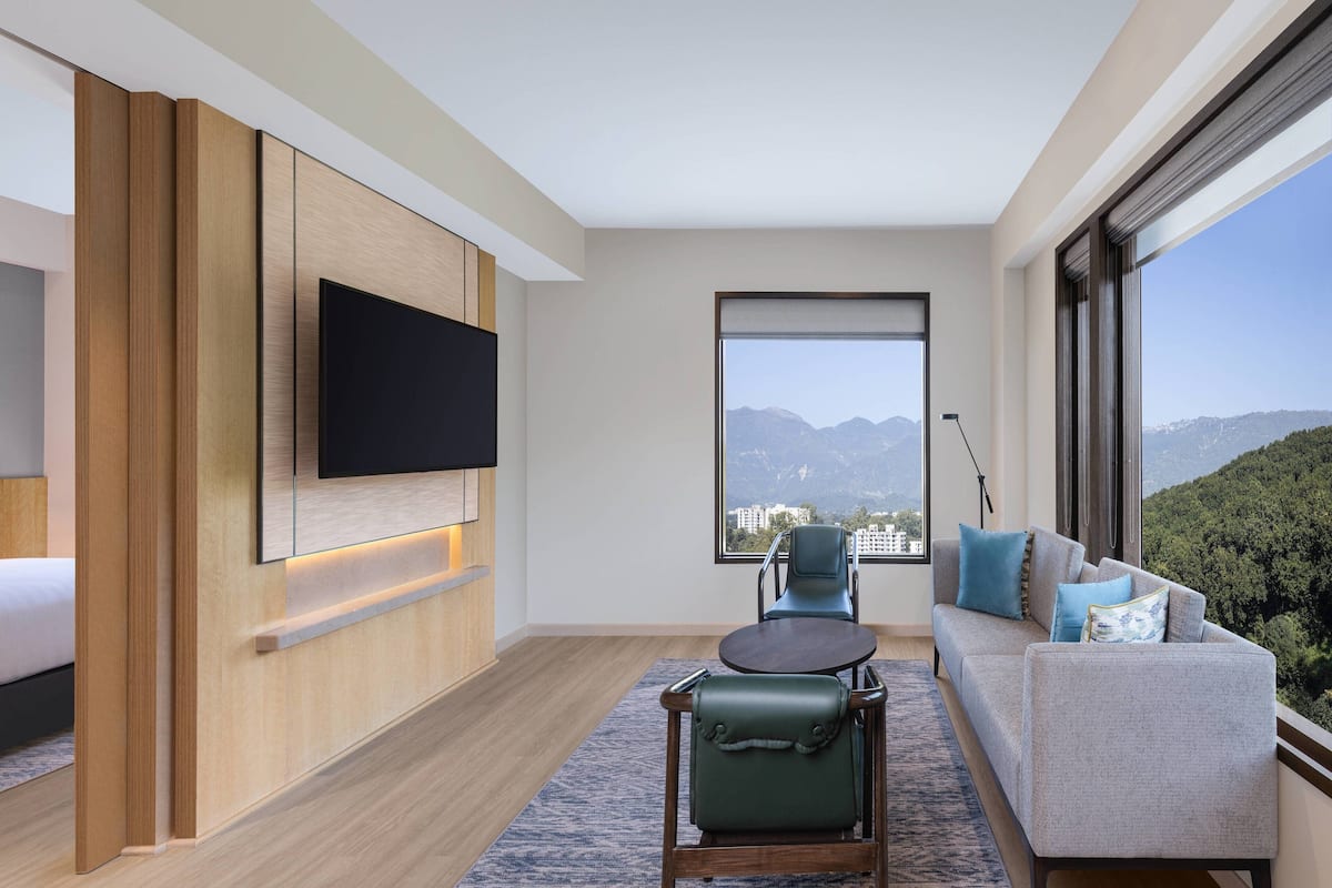 A modern hotel room with a large TV, a sofa, a chair, and a view of mountains.