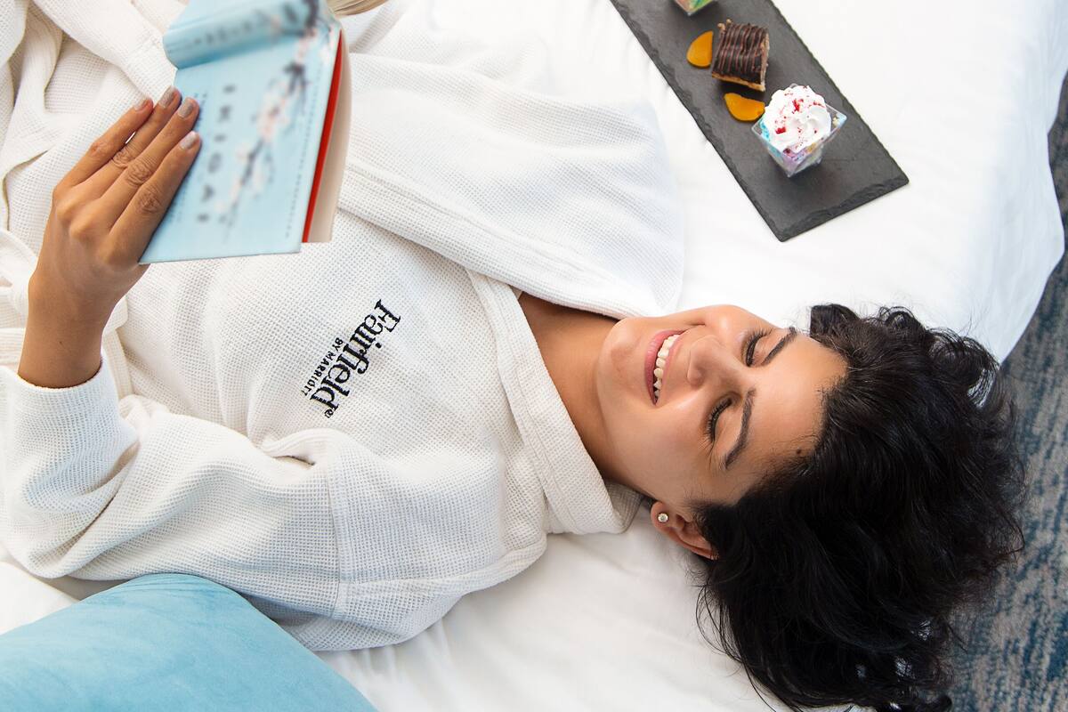 A person lying on a bed with a book, a plate of fruit, and a tray of pastries.