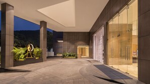 Entrada de un hotel moderno con fachada de vidrio, zona de recepción y espacio exterior ajardinado con una escultura.