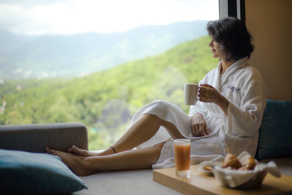 A person in a white robe sitting by a window with a view of green hills.