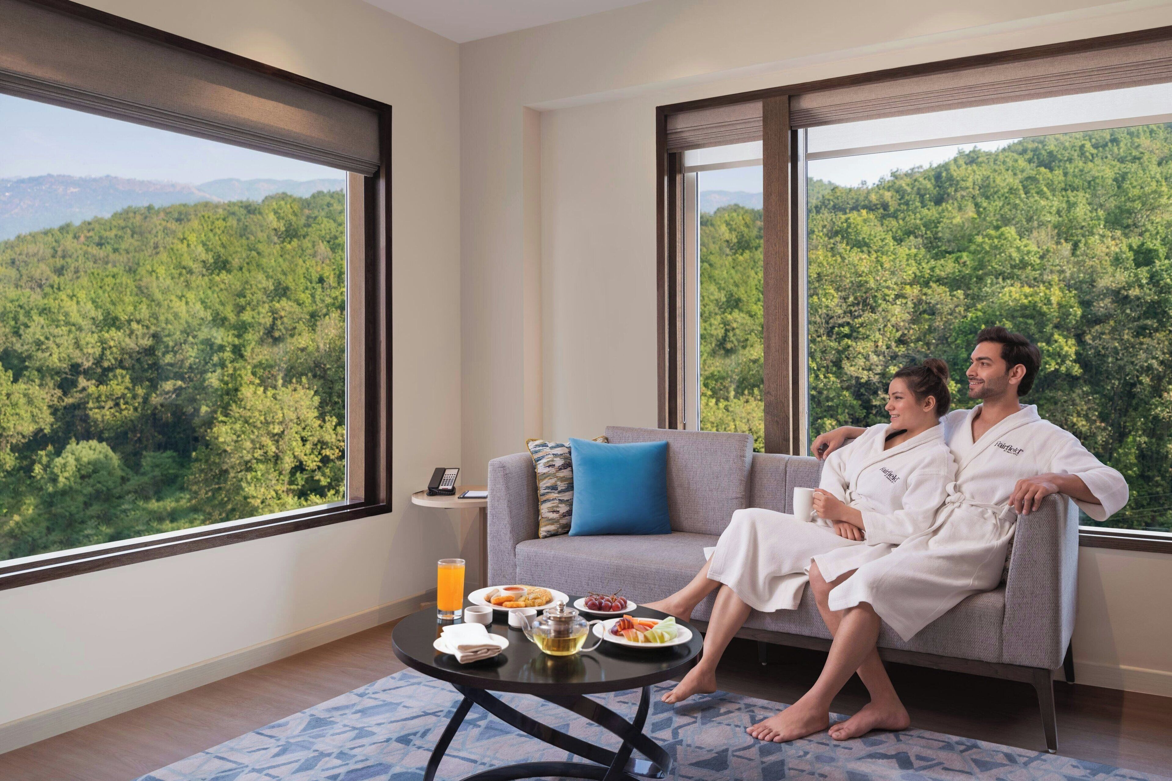 A room with a large window offering a forest view, a sofa with two people in robes, and a table with breakfast items.